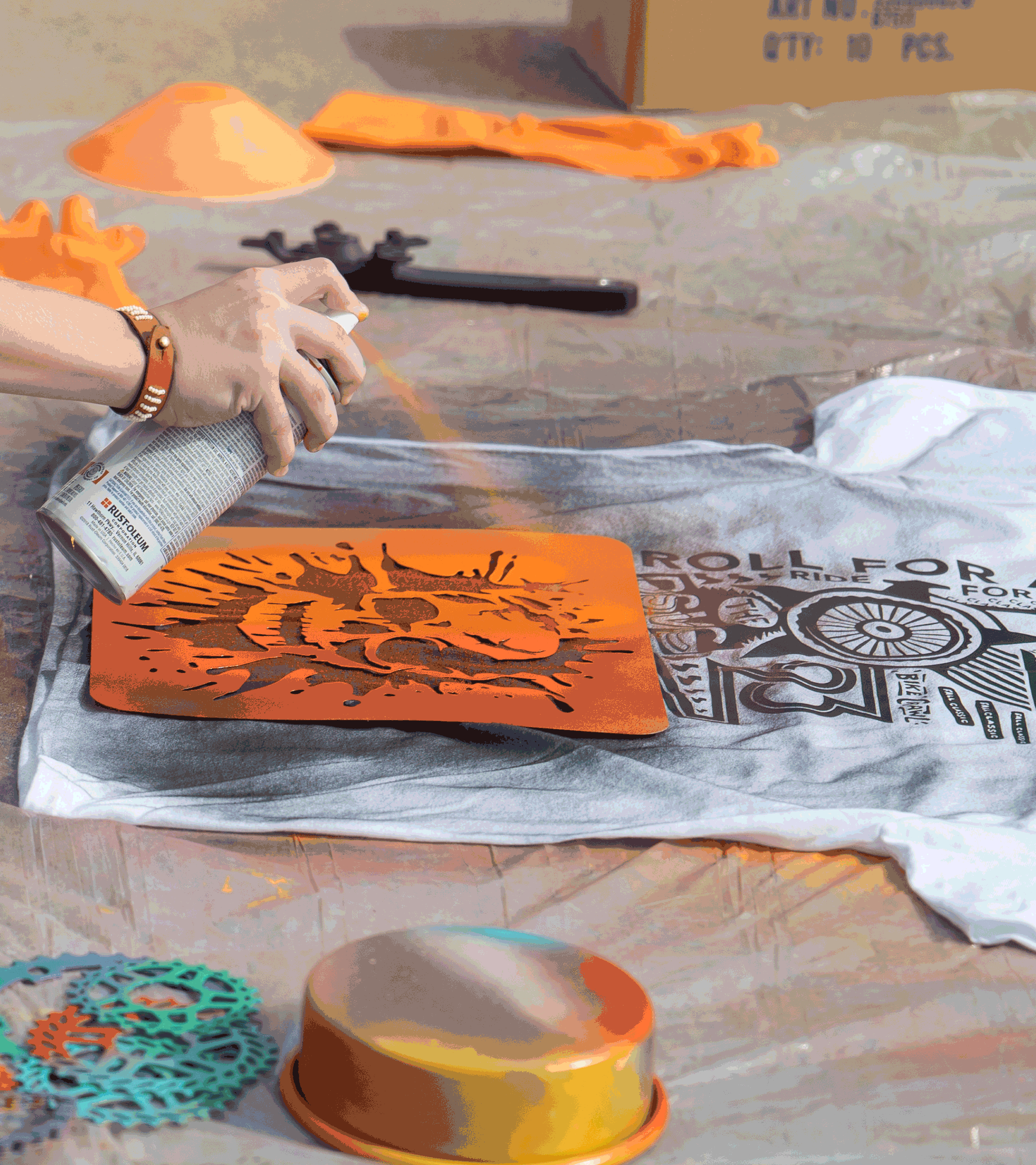 Image of a t-shirt being spray painted. A graphic of a skull is used as a stencil and the spray paint is orange.