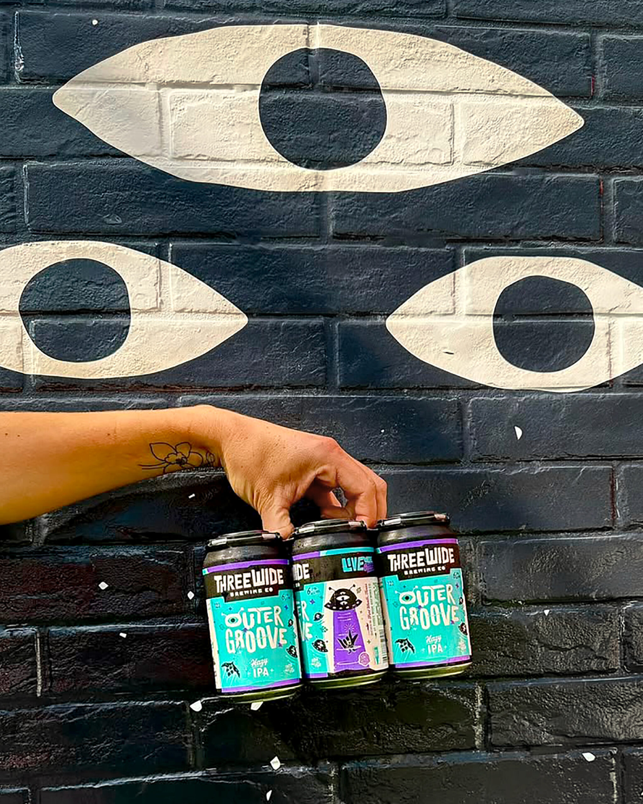 Photo of hand holding a pack of Three Wide "Outer Groove" beer in front of a brick wall with 3 eyes from the brand's characters. 